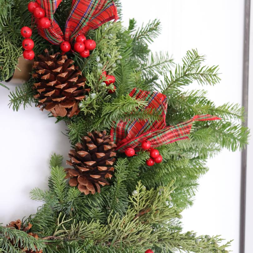 Gros plan sur des couronnes de Noël ornées de pommes de pin, de baies rouges et de ruban à carreaux sur fond blanc.