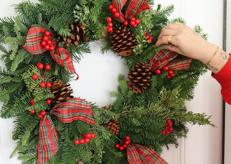Hand placing a decorative fresh Christmas wreath on a door with plaid ribbons and pinecones.