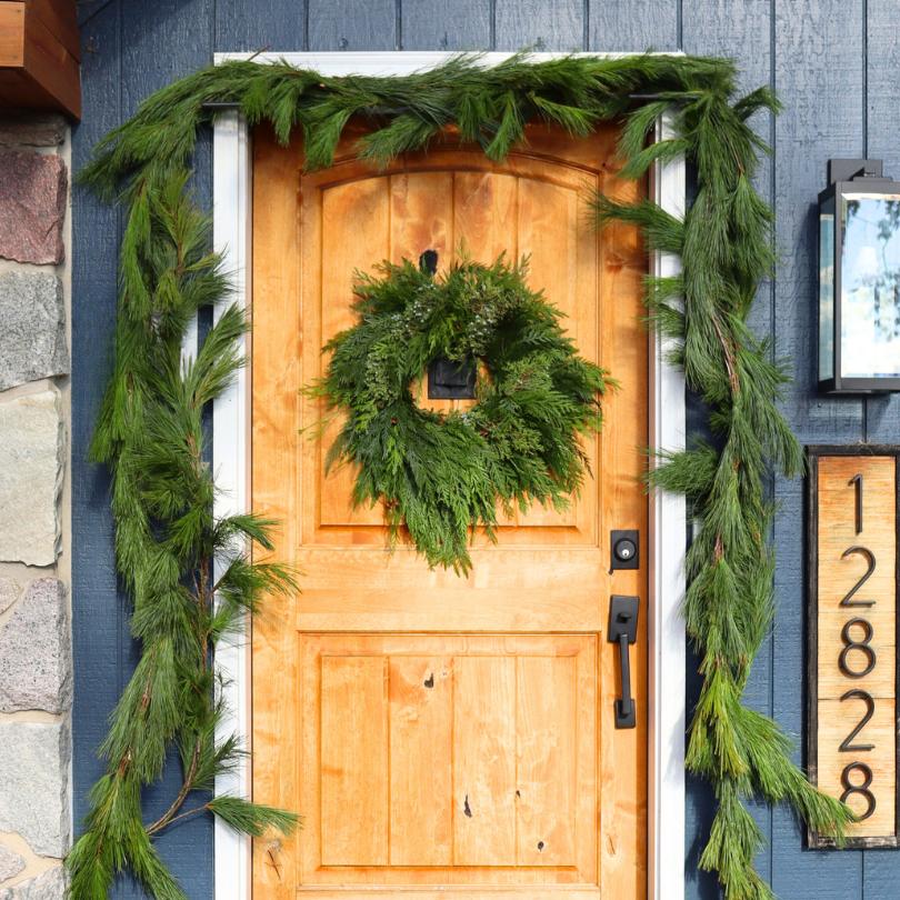 Porte en bois ornée d'une couronne de Noël en sapin et d'une guirlande de pin blanc frais, entourée de décorations festives sur la façade bleue d'une maison.