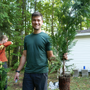 Man holding a 'Green Giant' arborvitae tree in a forested area with another person in the background.