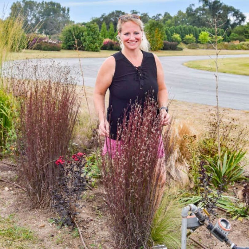 Une femme se tient debout dans un jardin avec de grands petits bluestems « Cinnamon Sticks » et une route en arrière-plan.